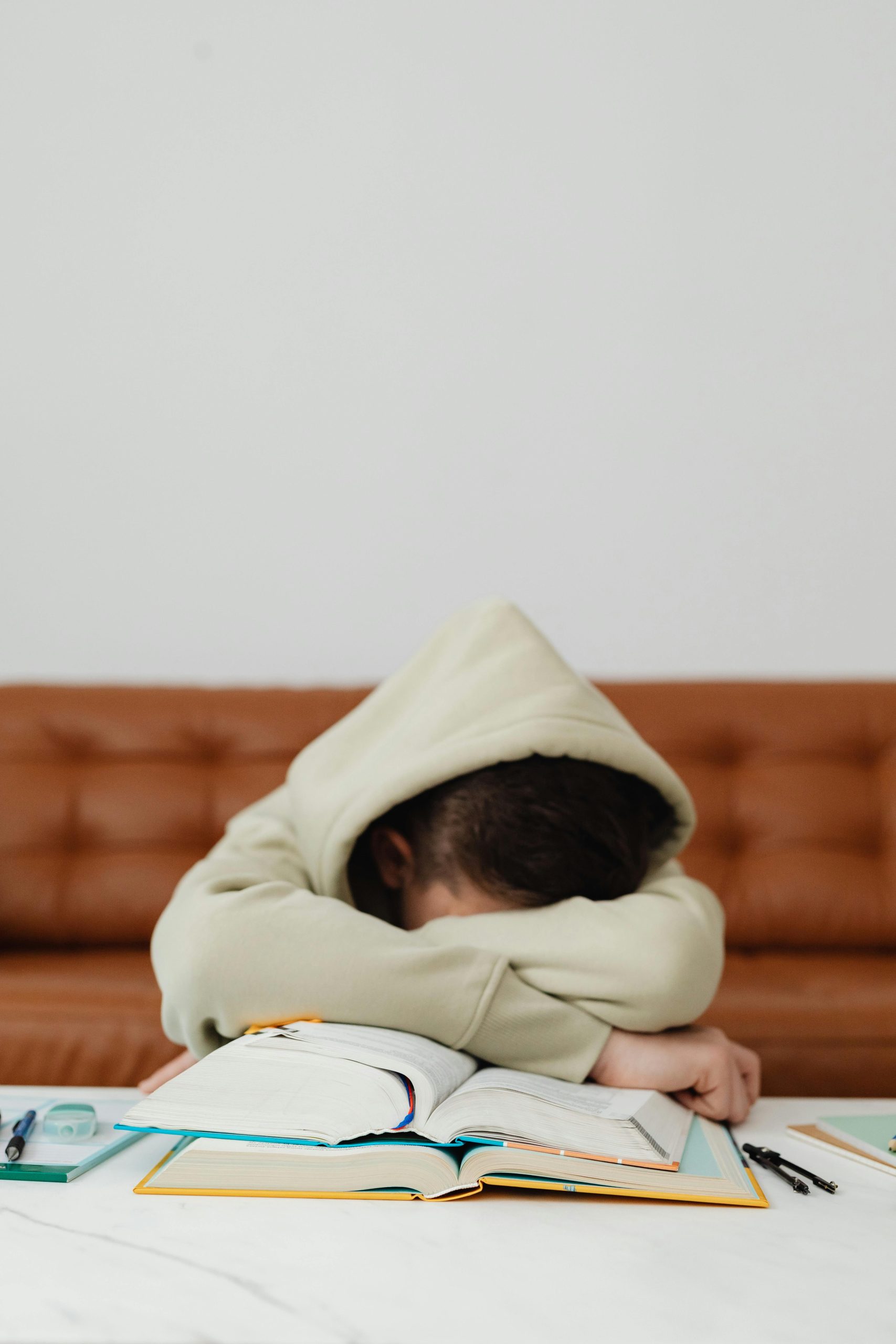 a child stressed with their hood up and their head on their arms on the table over their school books whilst studying