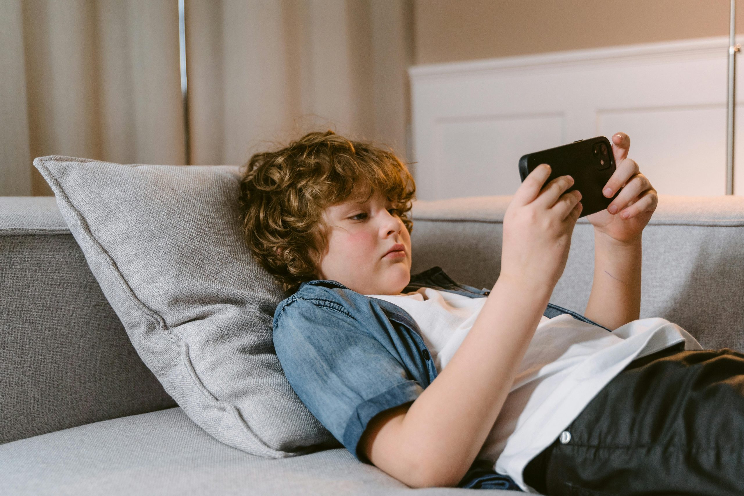 young boy lying slumped on the sofa looking low watching something on his smartphone