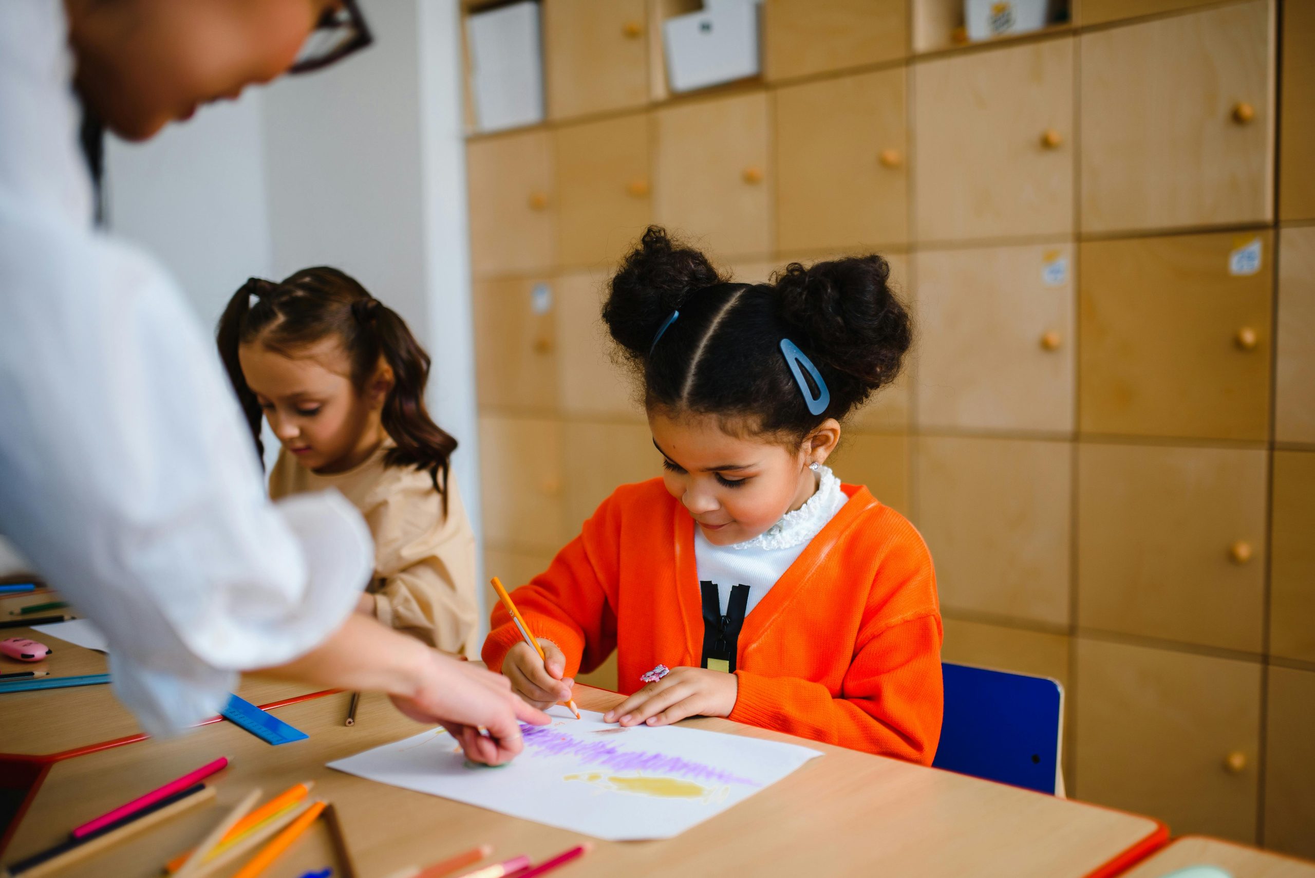 little girl drawing at primary school sat at her desk and a teacher helping her