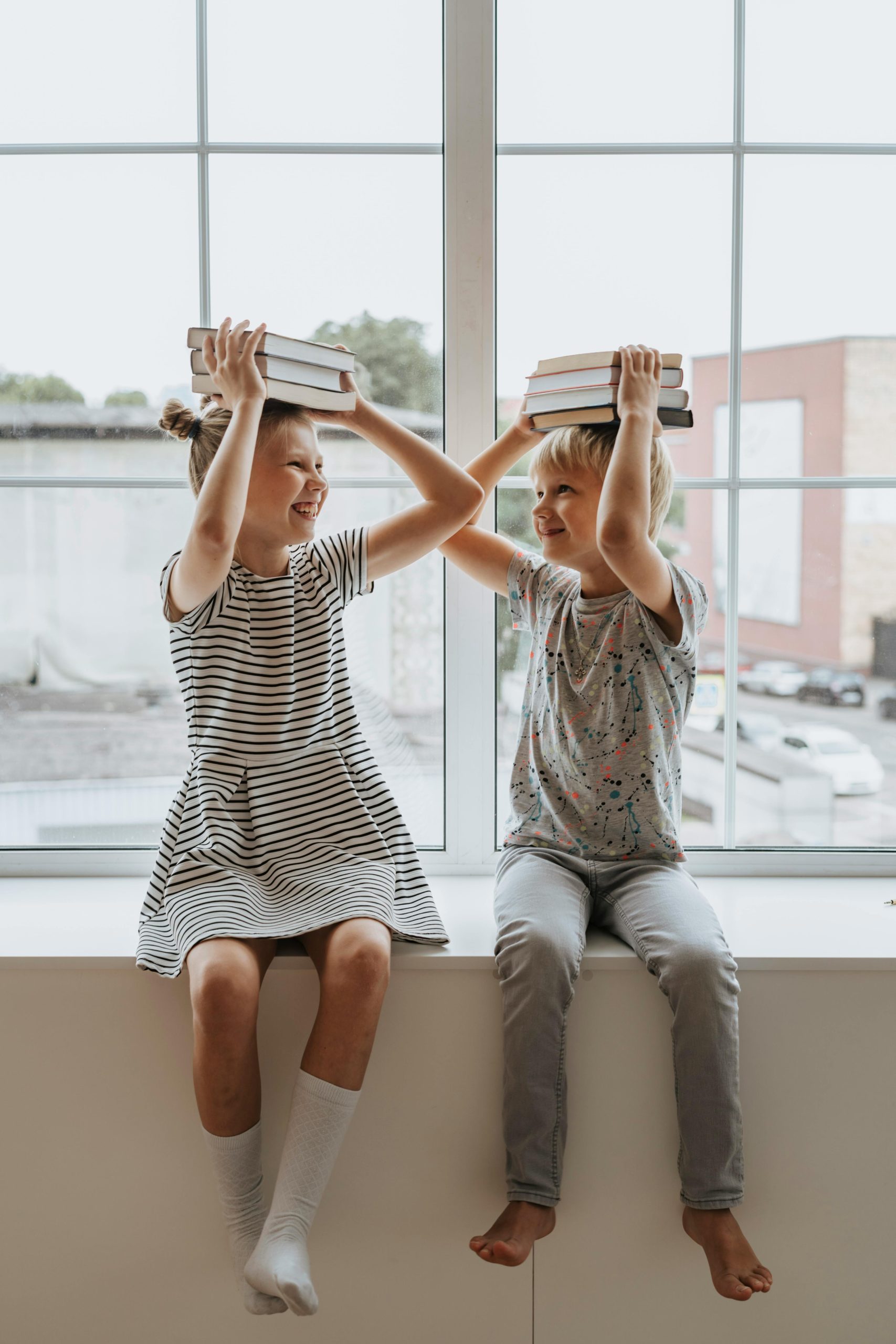 two children sat on a wall at school smiling holding books on their heads